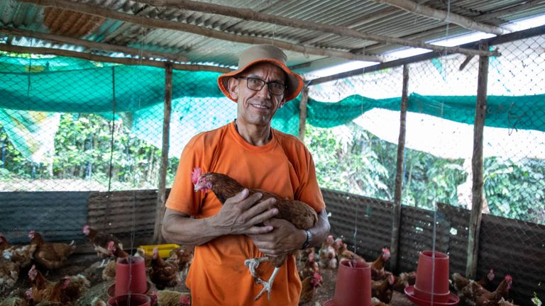 Smiling man in orange outfit holding a chicken in a rural coop.
